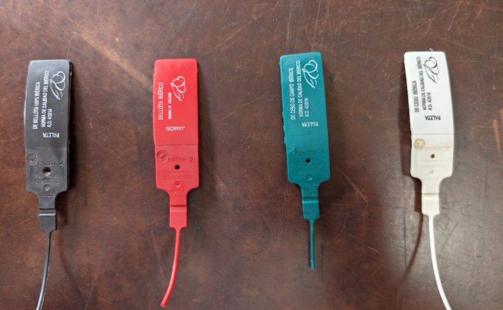 Color-coded Spanish Ibérico ham seals showing black, red, green, and white quality tags for different Jamón Ibérico categories.