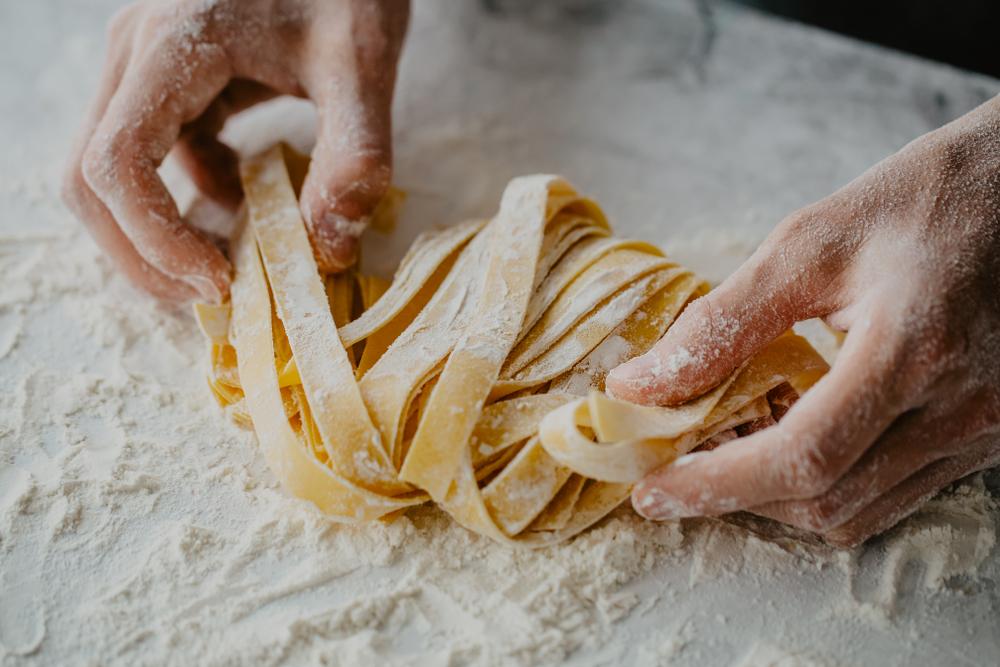 Hands shaping ribbons of fresh pasta on a floured surface.